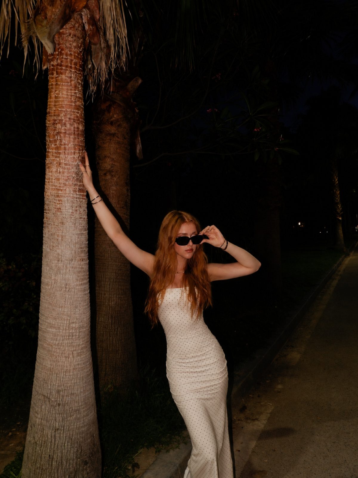 Delaney in a white polka-dot dress and sunglasses, leaning on a palm tree at night