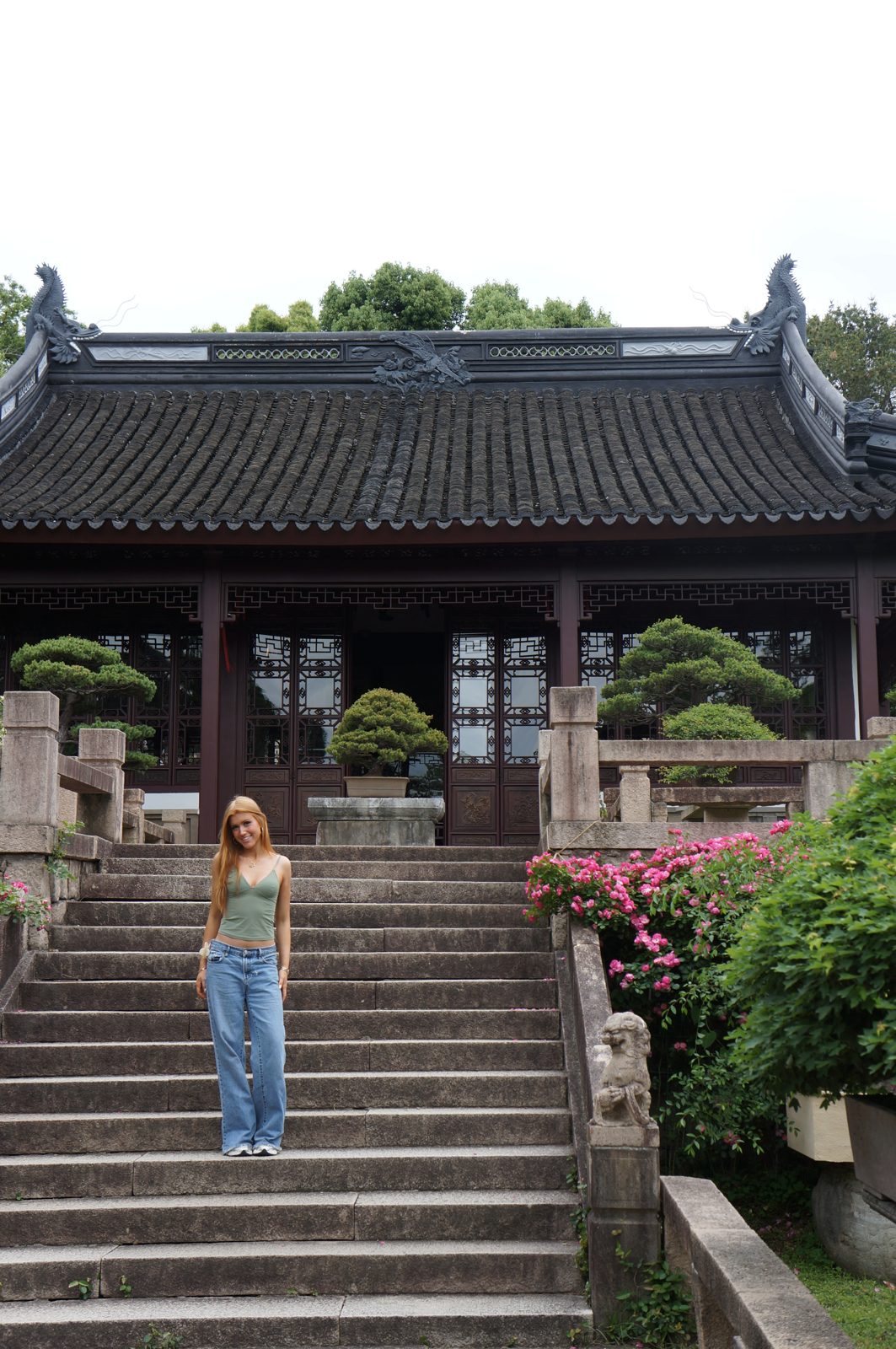 Delaney standing on stone steps in front of a traditional Chinese pavilion with bonsai trees and pink roses